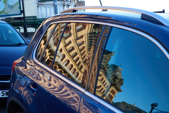 This urban photograph captures the reflection of the Scarborough Grand Hotel in the windows of a blue car parked on a street in Scarborough, Yorkshire. The image was taken in the late afternoon during the spring season, and the Grand Hotel’s distinctive architecture is clearly visible as it appears distorted and mirrored in the car's glass. The clear blue sky and natural lighting enhance the details of the hotel's ornate facade, showcasing one of the iconic landmarks of Scarborough in Yorkshire from an artistic perspective.